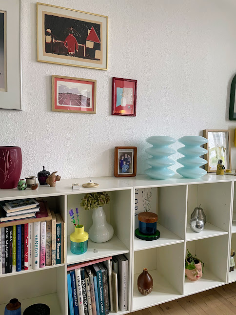 Hvid reol fyldt med kunstbøger, smukke glasvaser og collager på væggen